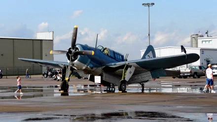 HD desktop wallpaper featuring a military aircraft parked on a wet tarmac with hangars and a few people in the background under a partly cloudy sky.
