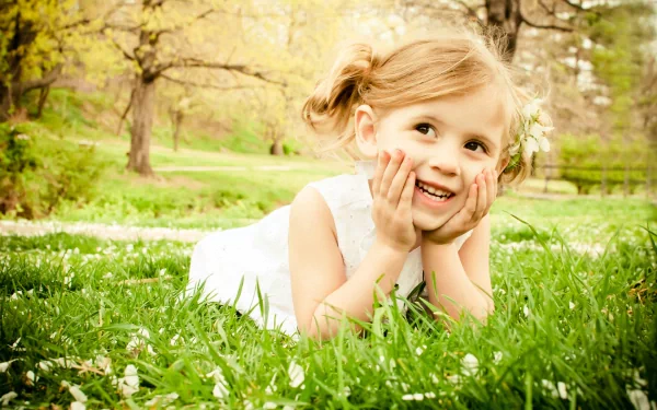 A cheerful child lying in grass, resting her chin on her hands, surrounded by blooming flowers and trees, captured in vibrant detail. A delightful HD desktop wallpaper.