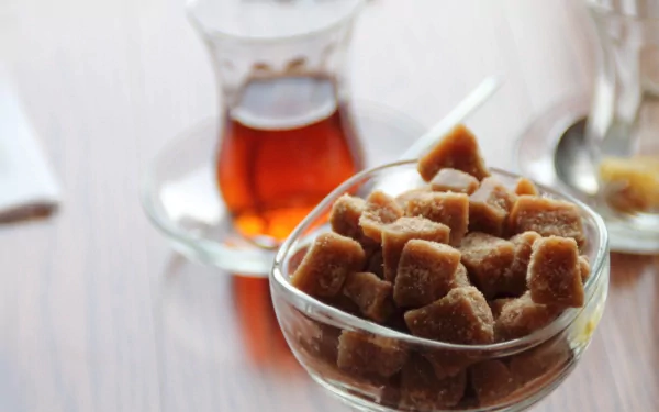 Close-up of brown sugar cubes in a glass bowl with a traditional tea glass filled with amber tea in the background, captured in vibrant 4K Ultra HD.