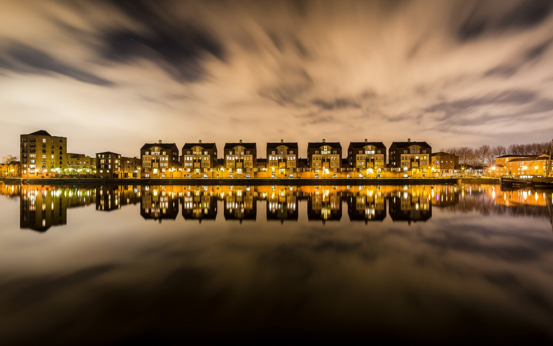 A stunning HD PC desktop wallpaper capturing city buildings illuminated at night, reflected perfectly on calm water under a dramatic, cloudy sky.