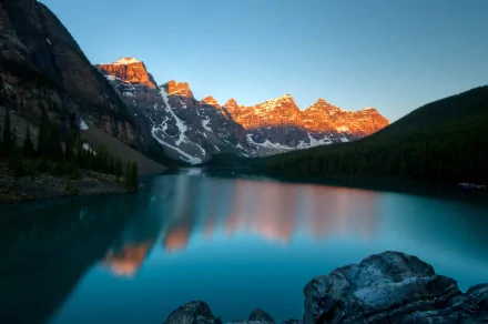 HD PC desktop wallpaper: golden sun on jagged mountain peaks reflecting in a turquoise alpine lake, rocky foreground and forested slopes under a clear blue sky.