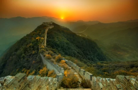 HD desktop wallpaper of the Great Wall of China at sunset, showcasing a stunning landscape with the sun setting behind mountains, casting an orange glow over the historic man-made structure.