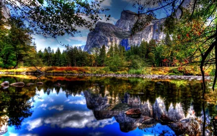 HD nature desktop wallpaper featuring a serene lake reflecting vibrant autumn trees and towering mountain cliffs under a blue sky with scattered clouds.