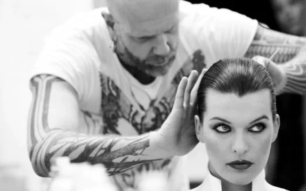 Black-and-white HD PC desktop wallpaper/background: close-up of a woman with slicked-back hair and dramatic eye makeup as a tattooed stylist adjusts her hairstyle.