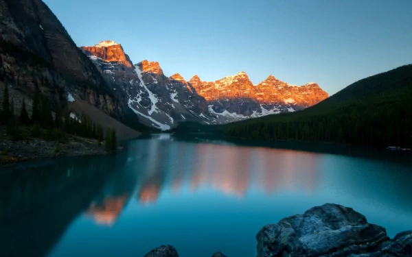 HD PC desktop wallpaper: golden sun on jagged mountain peaks reflecting in a turquoise alpine lake, rocky foreground and forested slopes under a clear blue sky.
