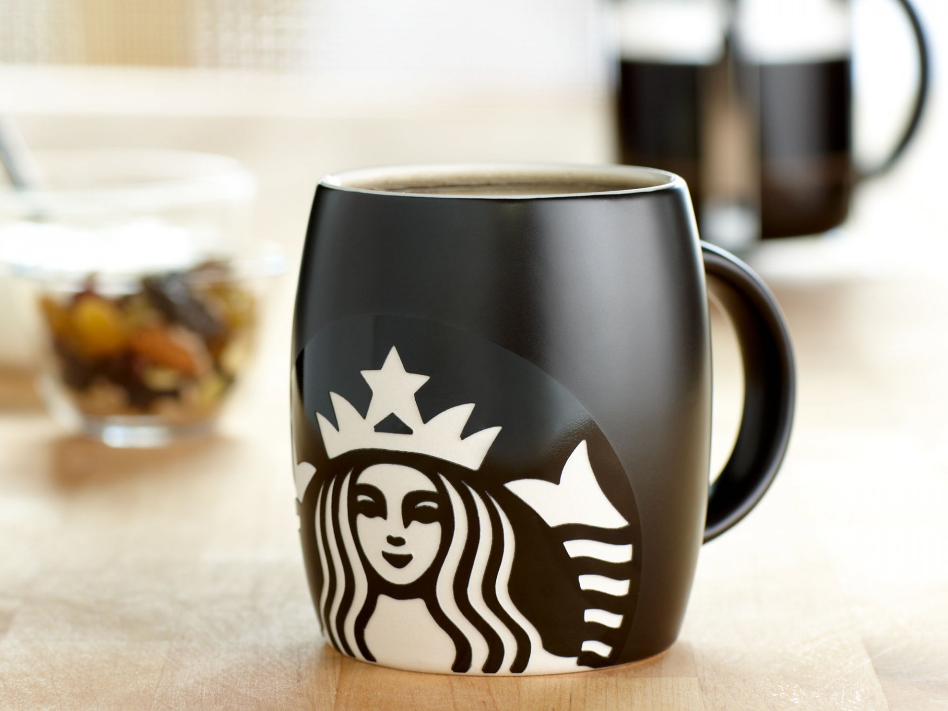 HD desktop wallpaper featuring a close-up of a black Starbucks coffee mug filled with beverage, highlighting the iconic white logo on a blurred kitchen background.