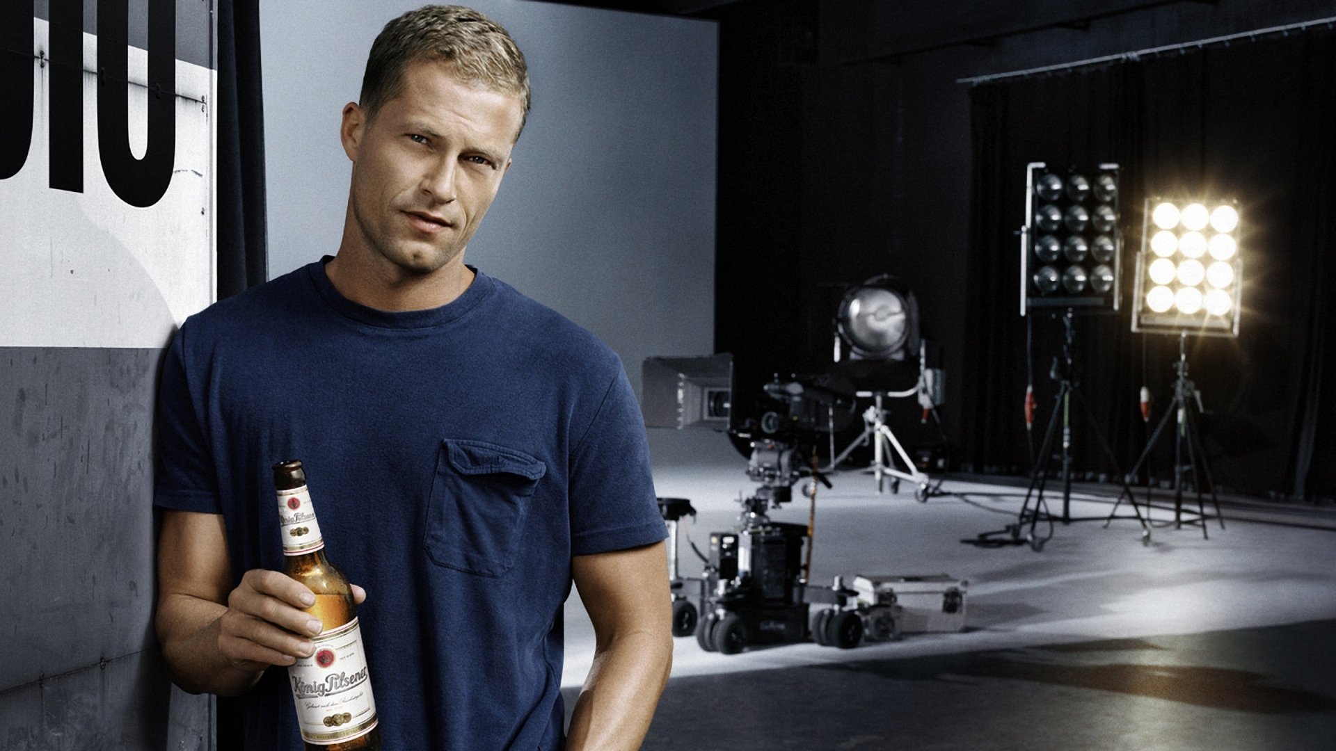 HD PC desktop wallpaper featuring celebrity Til Schweiger holding a beer bottle in a studio setting with professional lighting and camera equipment in the background.
