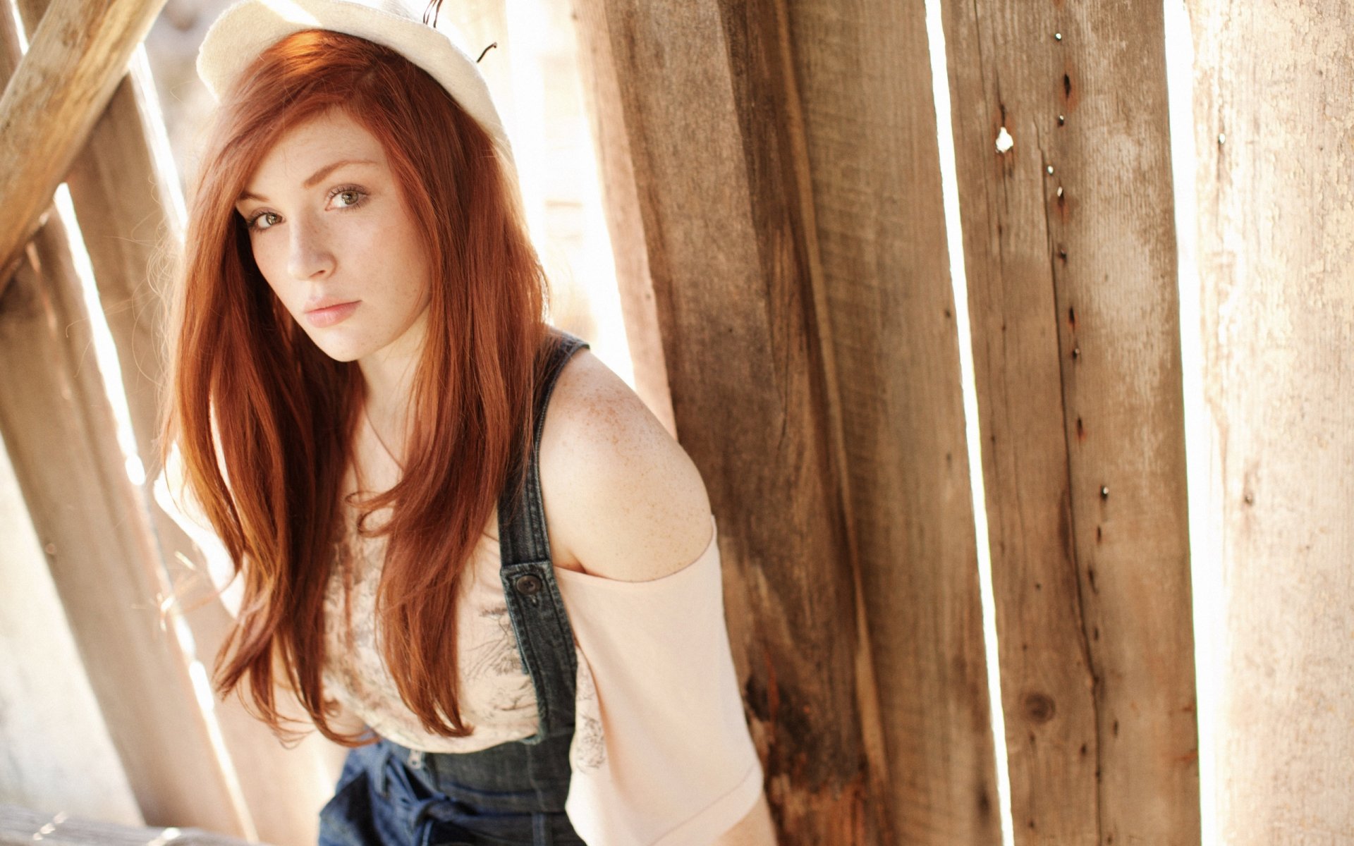 A young woman with long red hair sits against a wooden wall, wearing a white knit hat and a casual outfit, captured in a high-definition desktop wallpaper.