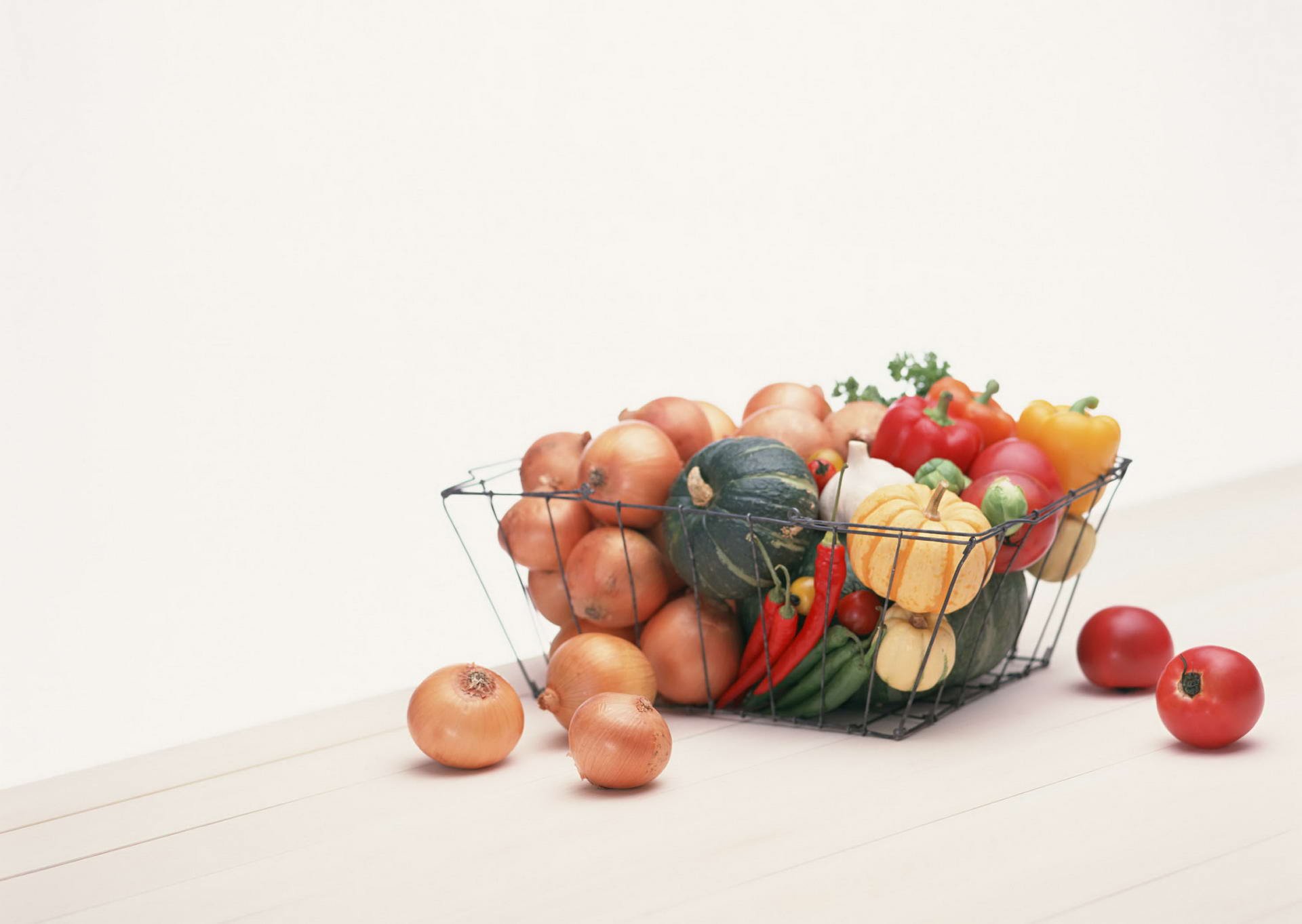HD desktop wallpaper featuring a wire basket filled with onions, tomatoes, peppers, and squash against a clean white background.