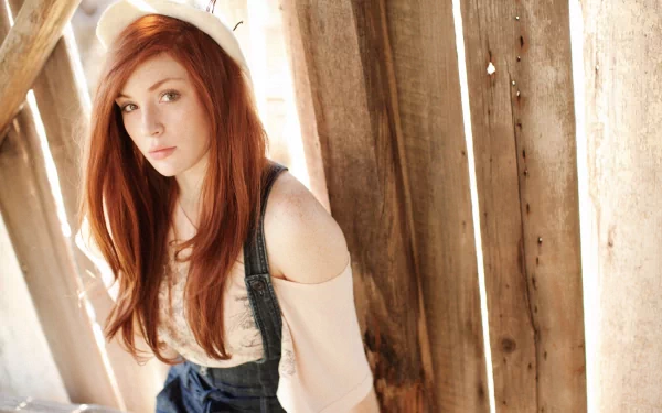A young woman with long red hair sits against a wooden wall, wearing a white knit hat and a casual outfit, captured in a high-definition desktop wallpaper.
