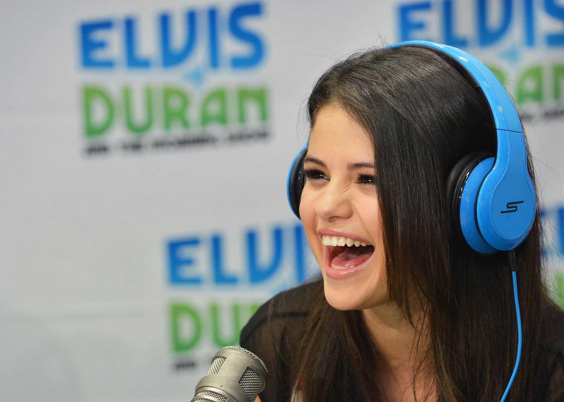 A vibrant HD desktop wallpaper featuring Selena Gomez smiling joyfully while wearing blue headphones, set against a backdrop of the Elvis Duran show logo.