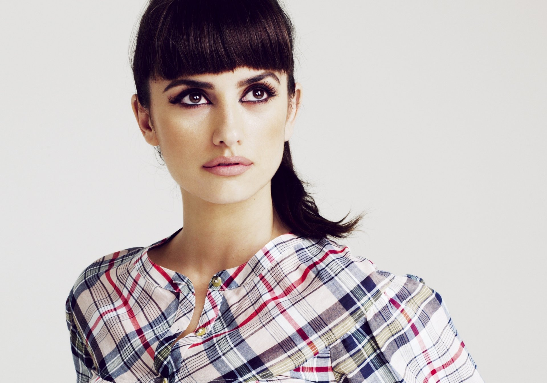 Penelope Cruz poses elegantly in a stylish, checkered top, showcasing her striking features and captivating gaze in this HD desktop wallpaper.