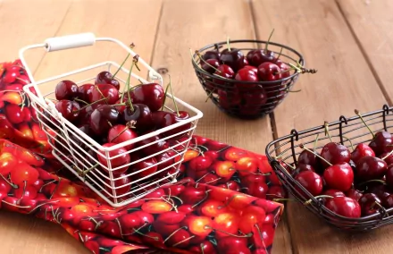 HD desktop wallpaper featuring fresh cherries in wire baskets on a wooden table with a cherry-patterned cloth beneath.