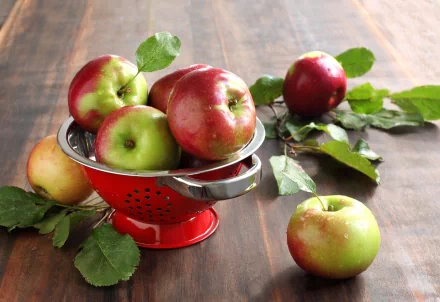 HD PC desktop wallpaper featuring fresh red and green apples in a red colander on a wooden surface with scattered green leaves.