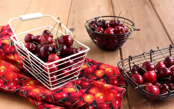 HD desktop wallpaper featuring fresh cherries in wire baskets on a wooden table with a cherry-patterned cloth beneath.