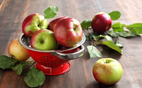 HD PC desktop wallpaper featuring fresh red and green apples in a red colander on a wooden surface with scattered green leaves.