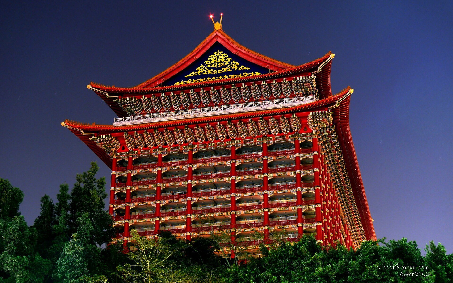 HD desktop wallpaper featuring a vibrant red religious pagoda illuminated against a deep blue evening sky, surrounded by lush greenery.