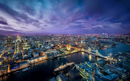 Aerial view of London's Tower Bridge at night, illuminated against the Thames River, showcasing a vibrant cityscape filled with lights and the bustling energy of the city.