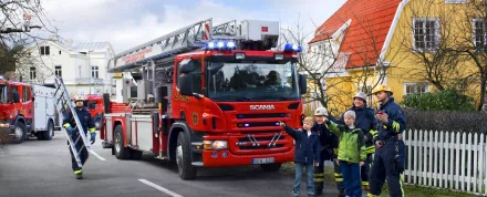 2K Quad HD PC desktop wallpaper of a red Scania fire truck (Scania fire engine vehicle) with firefighters and children waving along a street.