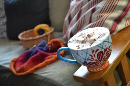 A cozy coffee scene featuring a beautifully decorated mug with frothy milk and chocolate sprinkles, set on a wooden surface amidst warm, colorful textiles.