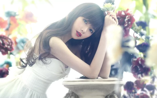 A woman with long brunette hair and red lipstick in a white dress leans gracefully against a pedestal surrounded by flowers. This HD desktop wallpaper captures a serene and elegant moment.