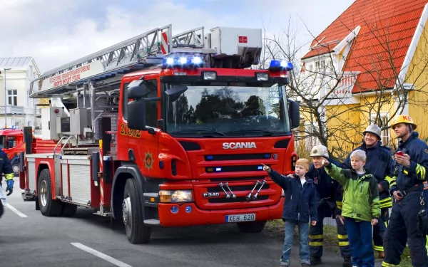 2K Quad HD PC desktop wallpaper of a red Scania fire truck (Scania fire engine vehicle) with firefighters and children waving along a street.