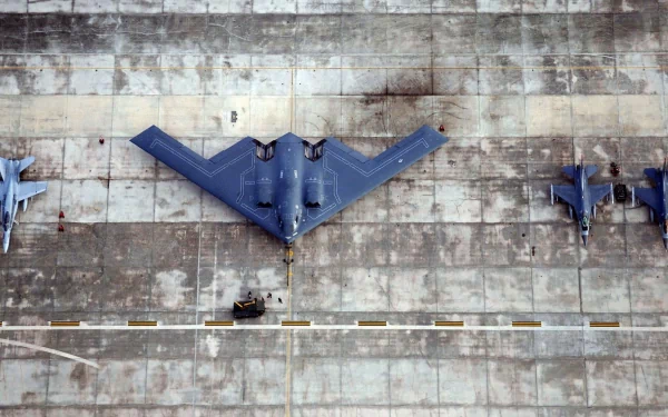 Top-down view of a Northrop B-2 Spirit stealth bomber on a concrete tarmac with other military aircraft — 2K Quad HD PC desktop wallpaper/background.