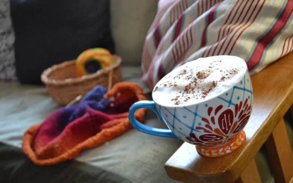 A cozy coffee scene featuring a beautifully decorated mug with frothy milk and chocolate sprinkles, set on a wooden surface amidst warm, colorful textiles.