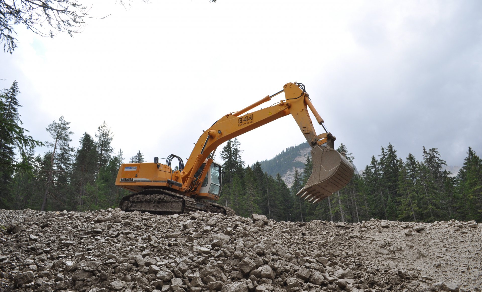 Yellow Liebherr 944 excavator vehicle on a rocky slope with bucket raised, forested mountains and cloudy sky backdrop — 4K Ultra HD PC desktop wallpaper/background.
