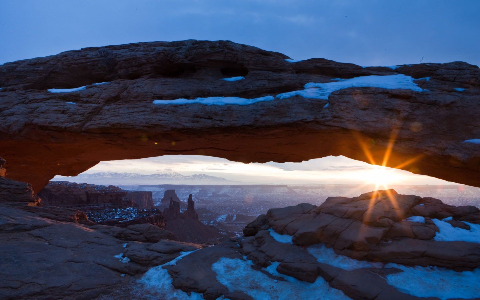 2K Quad HD PC desktop wallpaper/background: nature sunrise with a starburst through a sandstone arch, snow-dusted rocks and distant mesas beneath a blue dawn sky.