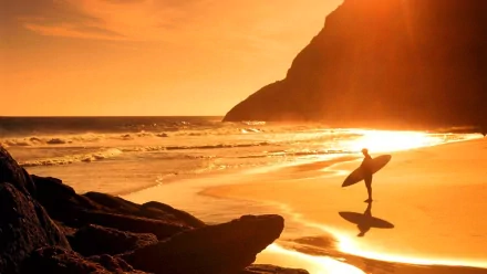 A surfer stands on a sunlit beach, holding a surfboard, with golden waves crashing in the background and rocky cliffs framing the scene. Perfect HD desktop wallpaper for surfing enthusiasts.