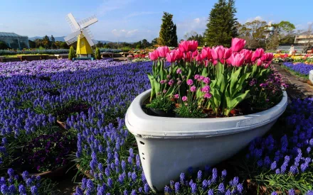 HD desktop wallpaper of a picturesque garden featuring vibrant pink and purple flowers in a boat-shaped planter, with a yellow windmill in the background.