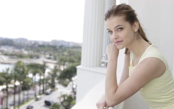 A stunning HD desktop wallpaper featuring celebrity Barbara Palvin, gazing thoughtfully while leaning on a balcony, with vibrant city scenery and palm trees in the background.