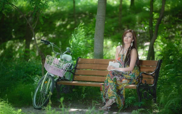 A woman sitting on a bench amidst lush greenery, holding a book with a bicycle nearby. She appears calm and thoughtful. This HD wallpaper captures a peaceful outdoor moment.
