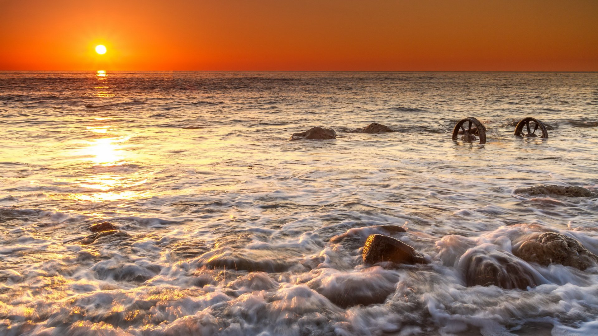 A stunning ocean sunset bathes the waves in golden light, with rocks and submerged wheels adding natural texture to this HD desktop wallpaper. Nature's beauty captured in serene motion.