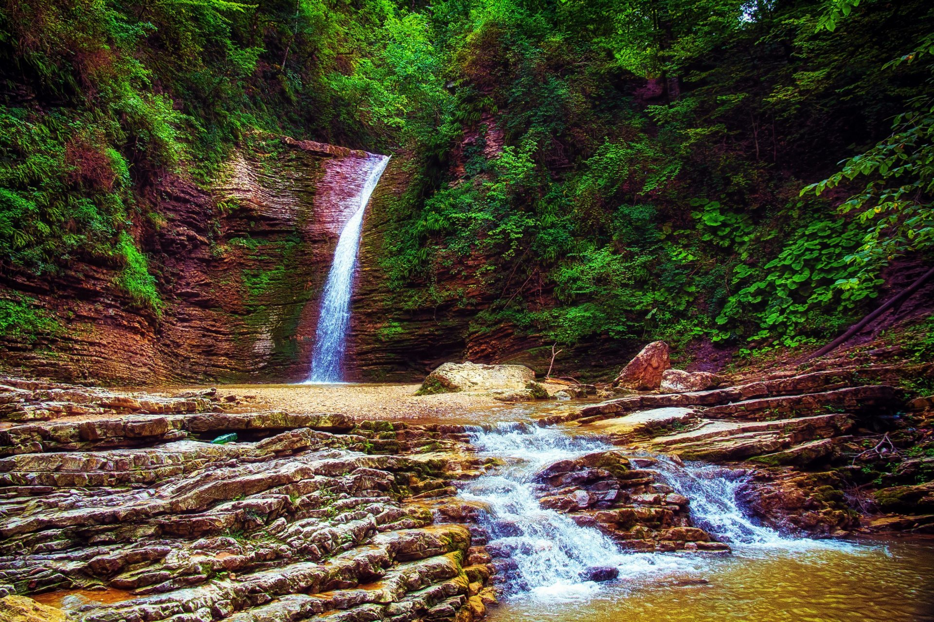 A serene waterfall cascades amidst lush greenery, surrounded by rocky terrain and a gentle stream, creating a tranquil scene that captures the essence of nature. HD desktop wallpaper.