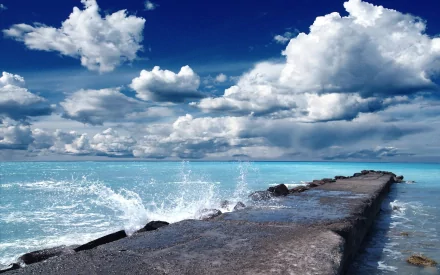 HD desktop wallpaper of a man-made pier extending into the ocean under a bright blue sky filled with scattered clouds. Waves splash against the pier's edge.