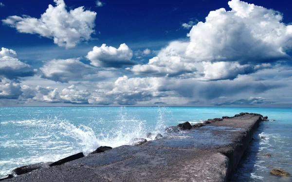 HD desktop wallpaper of a man-made pier extending into the ocean under a bright blue sky filled with scattered clouds. Waves splash against the pier's edge.