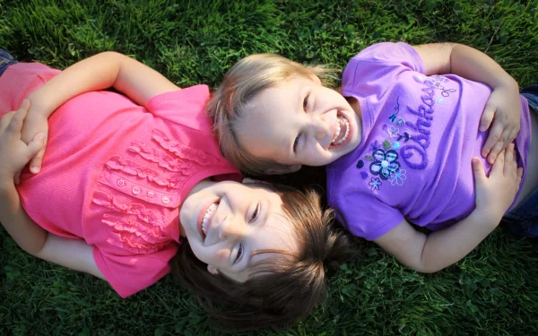 Two joyful children, lying on green grass, smiling and enjoying their time together, captured in vibrant HD photography. This image creates a cheerful and warm desktop wallpaper.