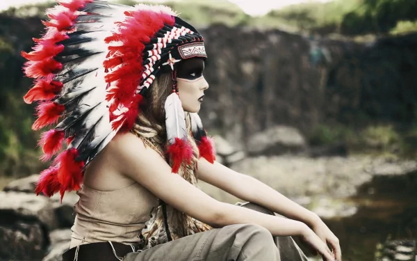 HD desktop wallpaper of a Native American woman wearing a traditional red and white feathered headdress, seated outdoors near rocky terrain.