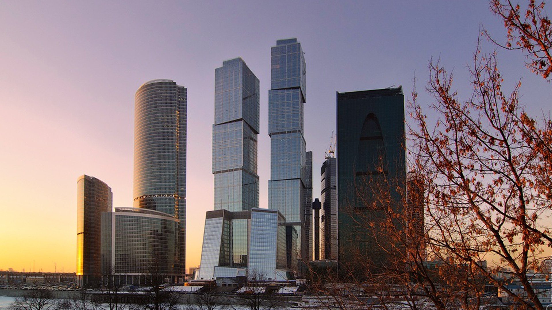 HD PC desktop wallpaper/background of man-made Moscow skyline at sunset, reflective modern skyscrapers rising behind snow and bare winter trees.