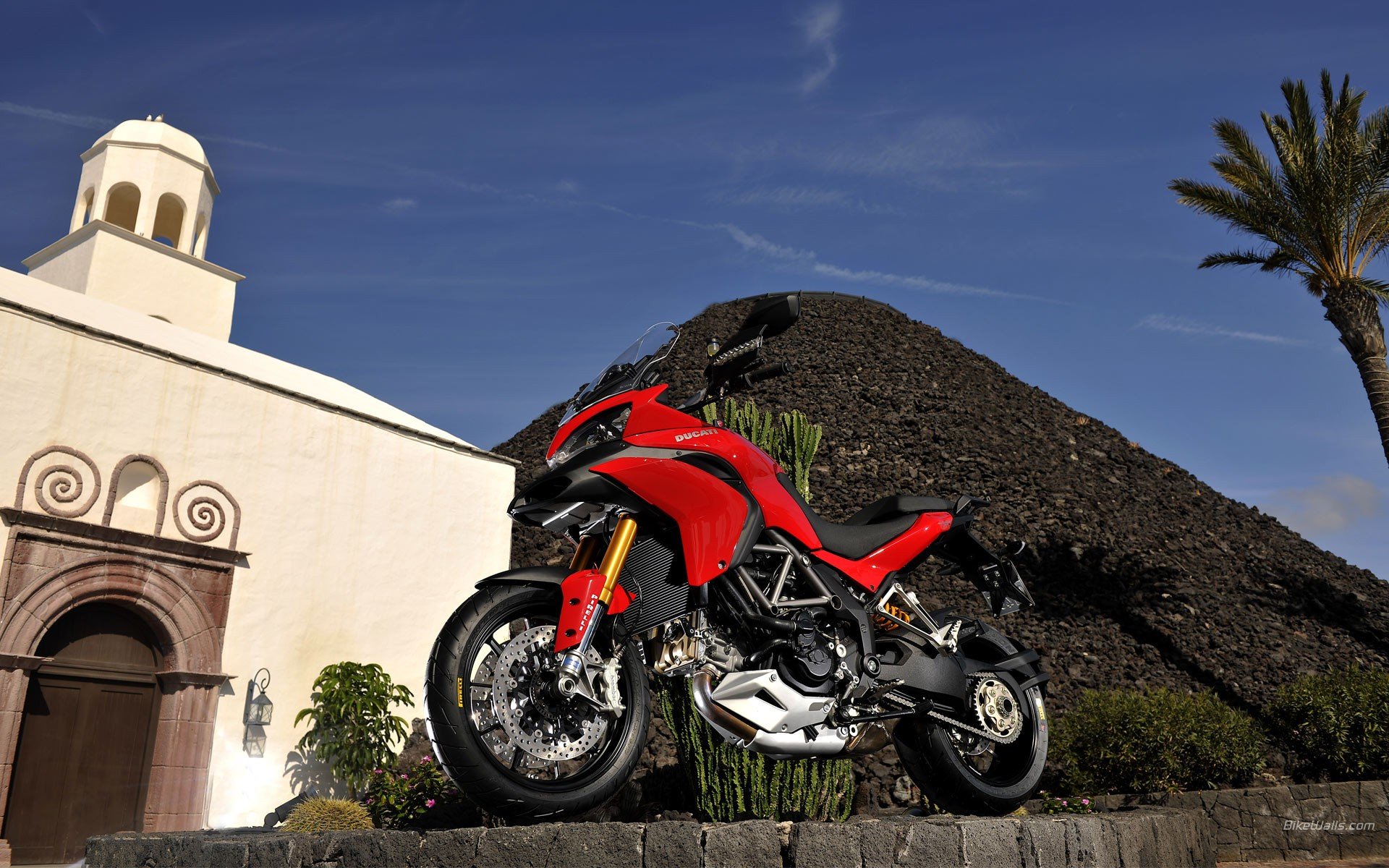HD PC desktop wallpaper featuring a red Ducati Multistrada 1200 motorcycle parked outdoors against a clear blue sky and historic architecture background.
