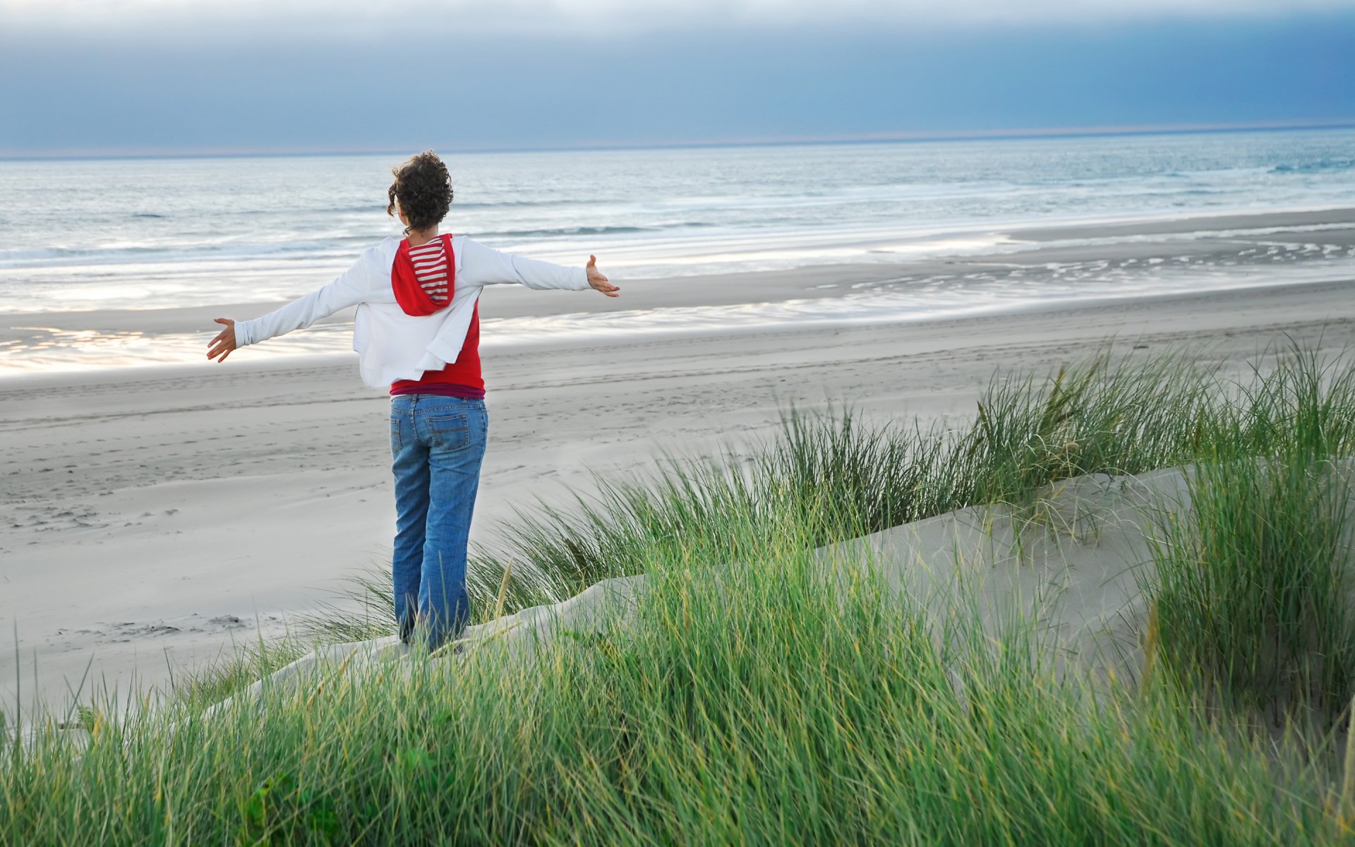HD PC desktop wallpaper of a woman standing on grassy dunes, arms outstretched, facing the calm ocean under a cloudy sky, evoking a serene and contemplative mood.