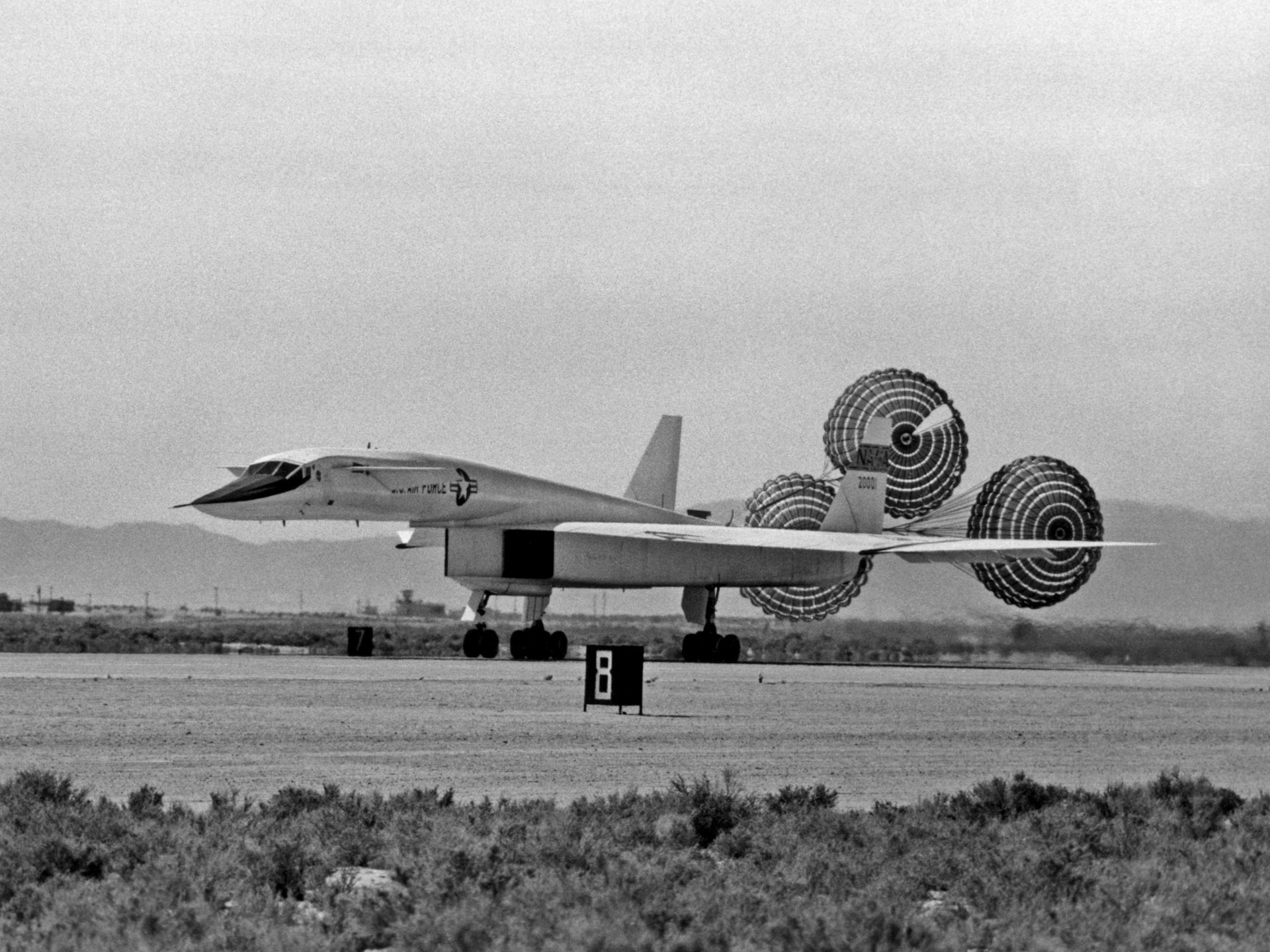 Black-and-white 2K Quad HD PC desktop background of a military North American XB-70 Valkyrie landing on a desert runway with its drag parachutes deployed.