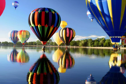 Hot air balloons in vibrant colors float over a calm lake, their reflections mirrored in the water. The image is an HD desktop wallpaper and background with clear skies and lush greenery.