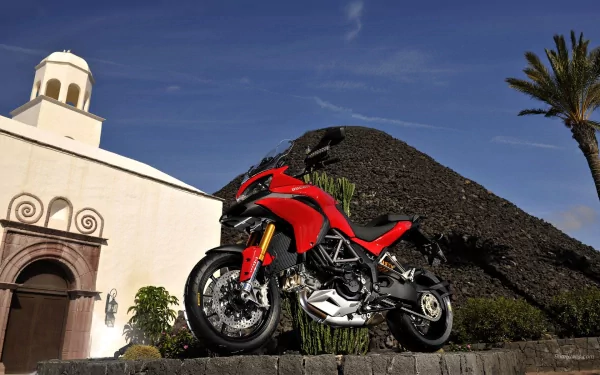 HD PC desktop wallpaper featuring a red Ducati Multistrada 1200 motorcycle parked outdoors against a clear blue sky and historic architecture background.