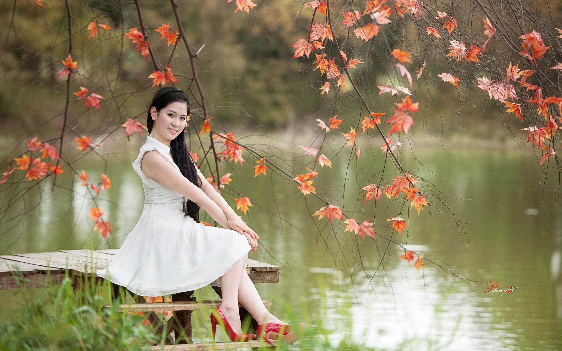 Asian woman in a white dress sitting on a bench by a tranquil lake, framed by autumn leaves. HD desktop wallpaper and background.