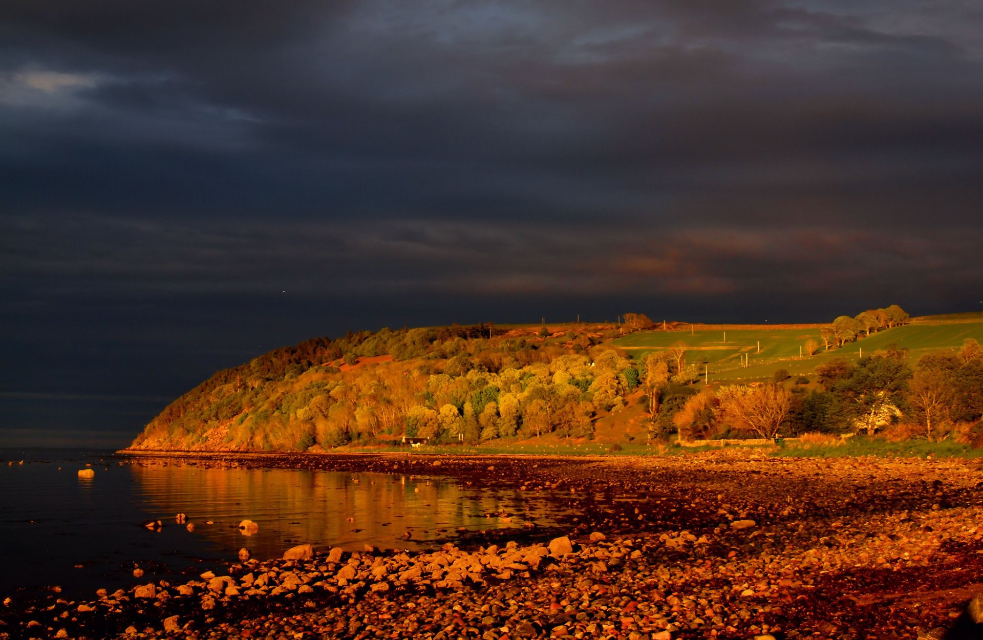 HD PC desktop wallpaper showcasing a serene lake with a rocky shore and a sunlit, tree-covered hill under a dramatic sky.