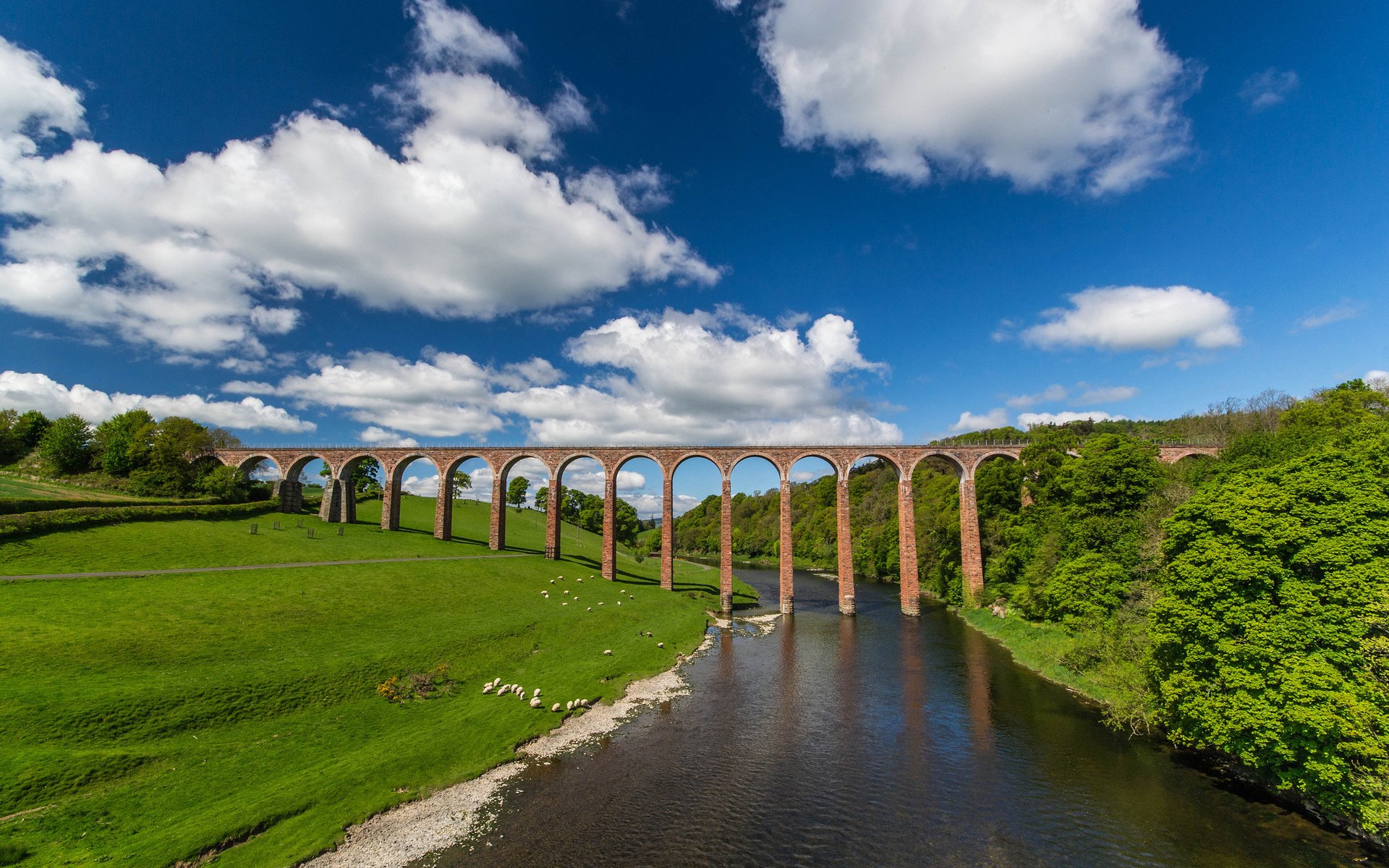A stunning HD wallpaper showcasing a majestic man-made bridge spanning a serene river, framed by lush green fields and a vibrant sky with fluffy clouds.