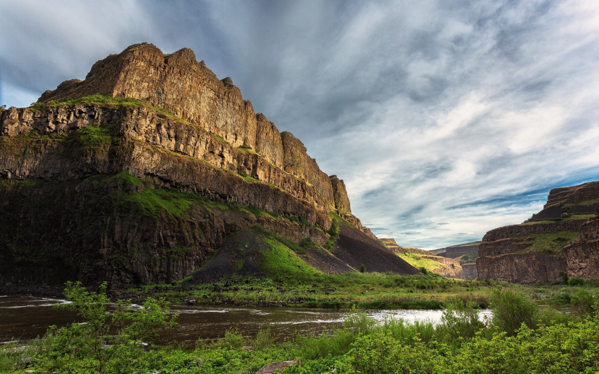 HD PC desktop wallpaper of a rugged nature cliff bathed in sunlight under a partly cloudy sky, with lush green vegetation and a winding river below.